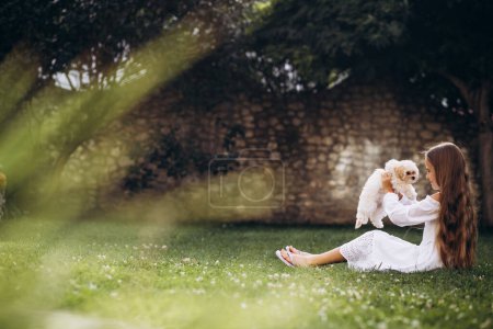 Téléchargez les photos : Petite fille avec son chien maltipa assis sur l'herbe dans la cour arrière de sa maison - en image libre de droit