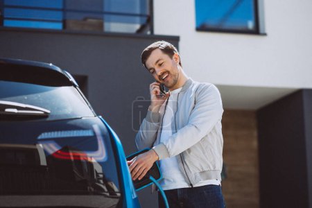 Photo for Young man charging electric car at home and using mobile phone - Royalty Free Image