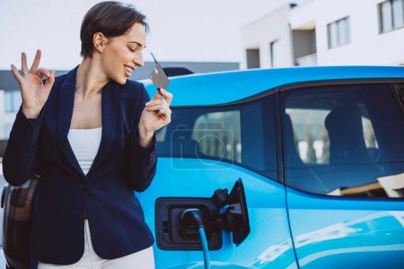Téléchargez les photos : Femme d'affaires debout près de sa voiture électrique tenant la carte de crédit - en image libre de droit