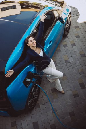 Foto de Mujer feliz de pie junto a su coche eléctrico - Imagen libre de derechos