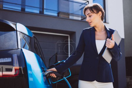 Foto de Mujer de negocios sosteniendo portátil por su coche eléctrico - Imagen libre de derechos