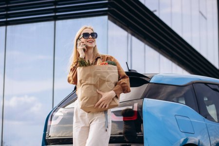 Foto de Mujer con bolsa de papel de supermercado junto a su coche eléctrico hablando por teléfono - Imagen libre de derechos