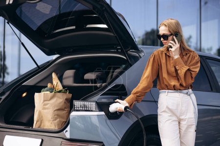 Photo for Woman with food shopping bags charging electric car and using mobile phone - Royalty Free Image