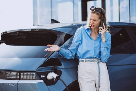 Photo for Business woman talking on the phone and standing by her electric car - Royalty Free Image