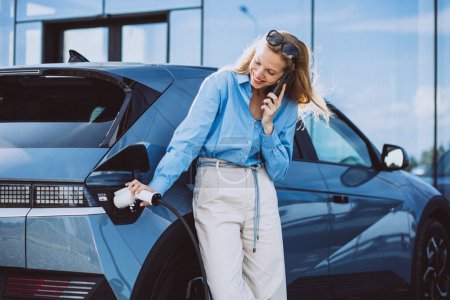 Photo for Business woman talking on the phone and standing by her electric car - Royalty Free Image