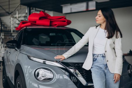 Téléchargez les photos : Femme dans la salle d'exposition de voiture debout à côté de sa nouvelle voiture décorée avec un énorme arc rouge - en image libre de droit