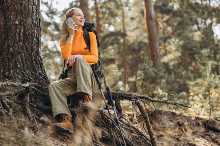 Téléchargez les photos : Femme voyageur avec sac et bâtons de marche assis près de l'arbre et en utilisant le téléphone - en image libre de droit