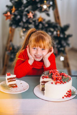 Foto de Niña de pelo rojo comiendo pastel de navidad junto al árbol de navidad - Imagen libre de derechos