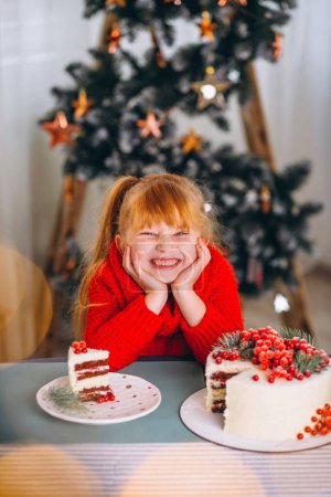 Foto de Niña de pelo rojo comiendo pastel de navidad junto al árbol de navidad - Imagen libre de derechos