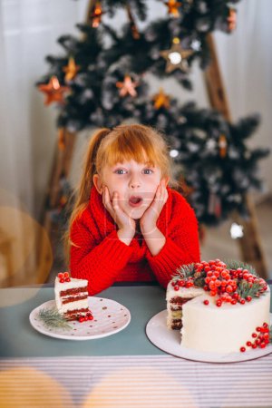 Téléchargez les photos : Petite fille aux cheveux roux mangeant un gâteau de Noël près de l'arbre de Noël - en image libre de droit