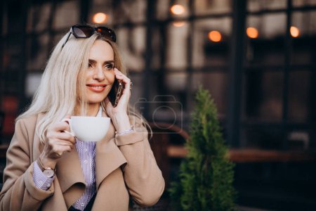 Photo for Elderly business woman drinking coffee outside cafe and talking on the phone - Royalty Free Image