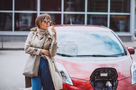 Foto de Mujer cargando electro coche en la gasolinera eléctrica y bebiendo café - Imagen libre de derechos
