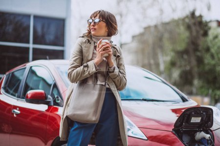 Téléchargez les photos : Femme charge electro voiture à la station-service électrique et boire du café - en image libre de droit