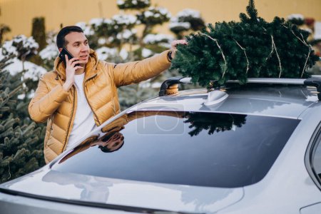 Photo for Handsome man talking on phone by the car with christmas tree on top - Royalty Free Image
