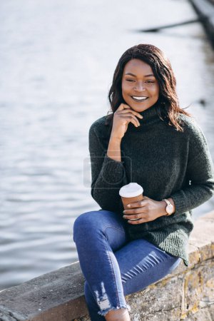 Téléchargez les photos : Jeune femme afro-américaine buvant du café au bord du lac - en image libre de droit