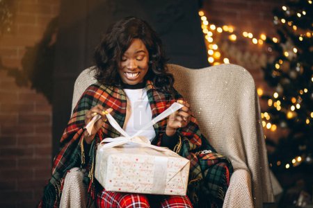 Téléchargez les photos : Femme afro-américaine assise sur une chaise et déballer des cadeaux de Noël - en image libre de droit