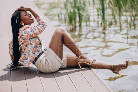 Téléchargez les photos : Élégante femme afro-américaine assise au bord du lac dans le parc - en image libre de droit