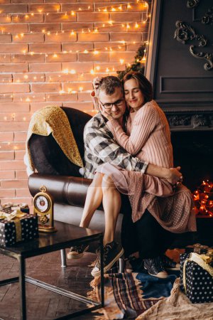Photo for Husband and wife sitting in chair by christmas tree - Royalty Free Image
