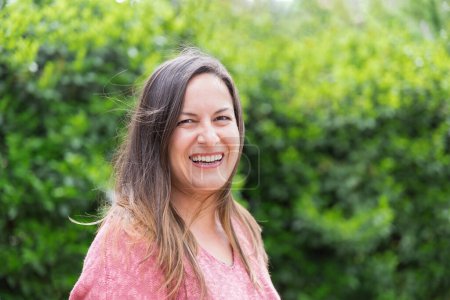 Outdoor portrait of a laughing 45 year old woman with windblown brown hair looking at the camera in the park on a sunny day. Confidence and tranquility of a mature woman. Happiness, lifestyle.