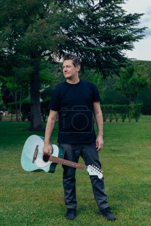 Photo for 40-50 year old man with tupe and gray hair posing with an acoustic guitar in a garden. Concept of learning and practicing a hobby - Royalty Free Image
