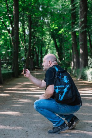 Photo for Portrait of a bald mature man with casual clothes and backpack taking a picture with his mobile phone in a park. Outdoor vertical portrait in natural light. - Royalty Free Image