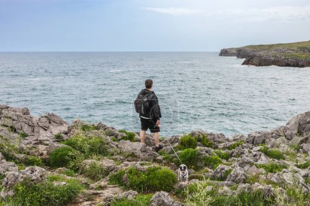 Photo for Back view of a mature man with backpack and casual clothes looking at the sea with his dog in nature. Travel and nature concept - Royalty Free Image