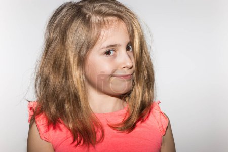 Téléchargez les photos : Portrait d'une petite fille très expressive de 8 ans aux cheveux clairs, t-shirt couleur saumon et regardant la caméra avec une expression désapprobatrice sur fond blanc. Concept d'enfant et d'enfance - en image libre de droit