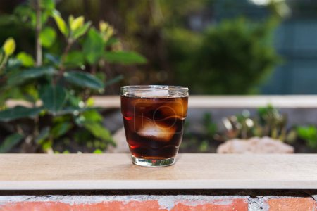 Photo for Front view of a glass filled with cola and ice, on a wallflower on an outdoor terrace on a summer evening. Summer concept, heat and refreshing drink - Royalty Free Image