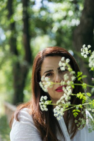 Photo for Portrait of a pretty 45 years old woman with red lips and looking at camera between flowers with green background of foliage in a park, on a sunny day. Concept of maturity, happiness and lifestyle. - Royalty Free Image