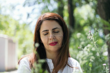 Photo for Portrait of a woman about 45 years old with red lips, relaxed and happy with closed eyes with green background of foliage on a sunny day in the park. Concept of maturity, happiness and lifestyle - Royalty Free Image