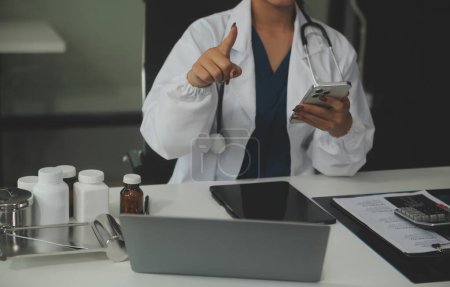 Seriöse Ärztin mit Laptop und Notizen in medizinischen Fachzeitschriften am Schreibtisch. Junge Ärztin mit weißem Mantel und Stethoskop arbeitet am Arbeitsplatz am Computer.