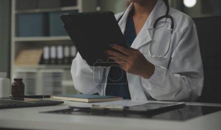 Serious female doctor using laptop and writing notes in medical journal sitting at desk. Young woman professional medic physician wearing white coat and stethoscope working on computer at workplace.