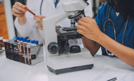 Foto de Trabajadora médica con bata de laboratorio y máscara estéril, haciendo un análisis de microscopio mientras su colega está trabajando detrás - Imagen libre de derechos