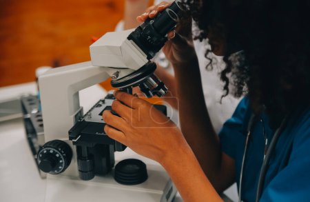 Foto de Trabajadora médica con bata de laboratorio y máscara estéril, haciendo un análisis de microscopio mientras su colega está trabajando detrás - Imagen libre de derechos