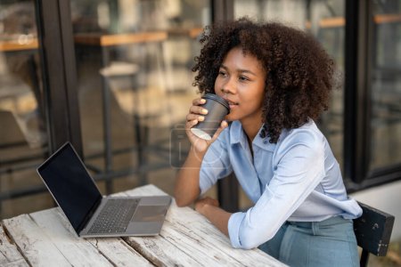 Téléchargez les photos : Jeune femme afro-américaine tenant tasse de café assis à l'extérieur du café avec ordinateur portable. Levé une tasse de café et bu et regardé loin. - en image libre de droit