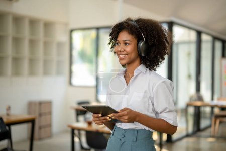 Téléchargez les photos : Étudiant afro-américain avec écouteurs à la main avec tablette debout souriant impatient dans un café ou un espace de travail - en image libre de droit