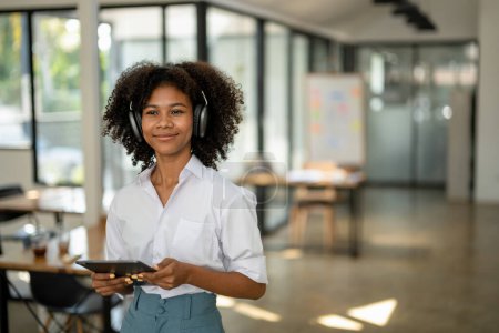 Téléchargez les photos : Étudiant afro-américain avec écouteurs à la main avec tablette debout souriant impatient dans un café ou un espace de travail - en image libre de droit
