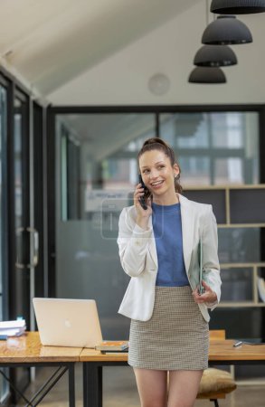 Foto de Mujer de negocios inteligente se para sosteniendo documentos y hablando por teléfono en la mesa de la oficina. - Imagen libre de derechos