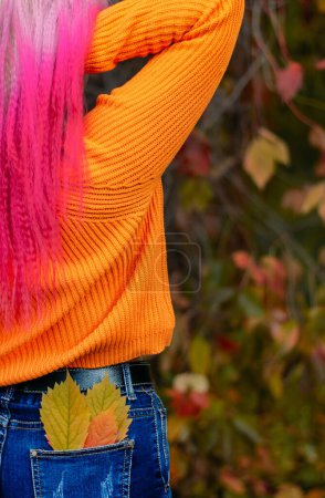 Foto de Una mujer de pelo rosado está de espaldas en un bosque otoñal, hojas rojas y amarillas sobresaliendo de su ropa. Explorando la naturaleza del otoño dorado. - Imagen libre de derechos
