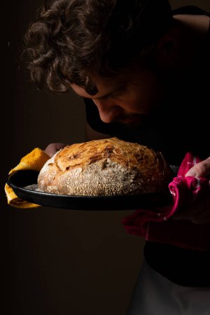 Téléchargez les photos : Joli jeune chef caucasien posant avec du pain au levain blanc. Le pain au levain est le protagoniste central de la scène, se détachant avec de belles tons dorés sur le fond sombre. - en image libre de droit