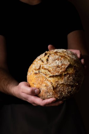 Téléchargez les photos : Joli jeune chef caucasien posant avec du pain au levain blanc. Le pain au levain est le protagoniste central de la scène, se détachant avec de belles tons dorés sur le fond sombre. - en image libre de droit