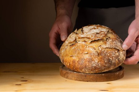 Téléchargez les photos : Les mains d'un jeune homme manipulant du pain au levain, mettant en valeur le pain avec de beaux tons dorés sur le fond sombre. - en image libre de droit