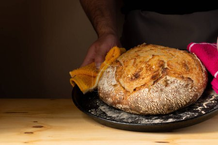 Téléchargez les photos : Les mains d'un jeune homme manipulant du pain au levain, mettant en valeur le pain avec de beaux tons dorés sur le fond sombre. - en image libre de droit