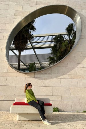 Foto de Mujer joven sentada en el banco frente a un edificio moderno. - Imagen libre de derechos