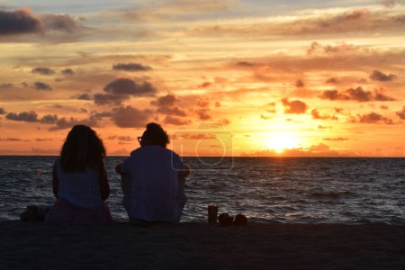 Téléchargez les photos : Jeune couple - homme et femme assis sur une plage de sable vide au coucher du soleil - en image libre de droit