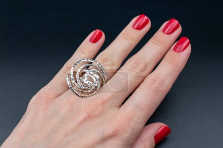 Female hand with red manicure holding silver ring on black background.