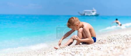 Téléchargez les photos : Un garçon joue avec des cailloux sur la belle plage de galets près de l'eau turquoise - en image libre de droit