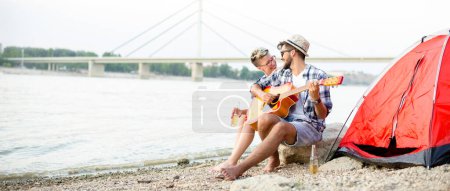 Boy is playing guitar to his girlfriend in camp on beach, panorama view