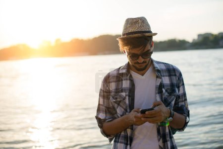 Junger Mann tippt auf seinem Smartphone am Strand, im Sonnenuntergang