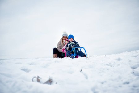 Mother with little children on holiday, preparing for tobogganing on snow covered mountain.